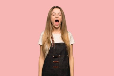 Young woman with apron shouting to the front with mouth wide open on isolated pink background