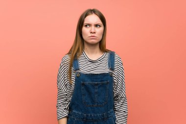 Üzücü ve depresif ifadesiyle pembe duvarın üzerinde tulumlu genç bir kadın.