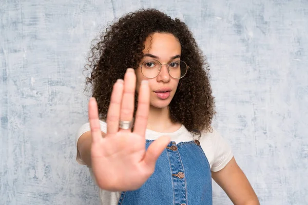 Dominican woman with overalls over grunge wall making stop gesture with her hand