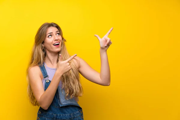 Mladá Blondýnka Montérkami Přes Izolované Žluté Pozadí Ukazuje Ukazováčkem Skvělý — Stock fotografie