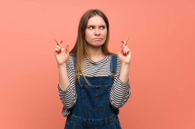 Pembe duvarda tulumlu, parmakları çapraz genç bir kadın.