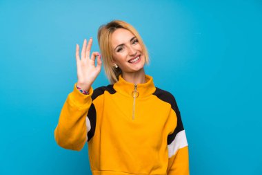 İzole edilmiş mavi arka planda genç sarışın kadın parmaklarıyla iyi bir işaret gösteriyor.