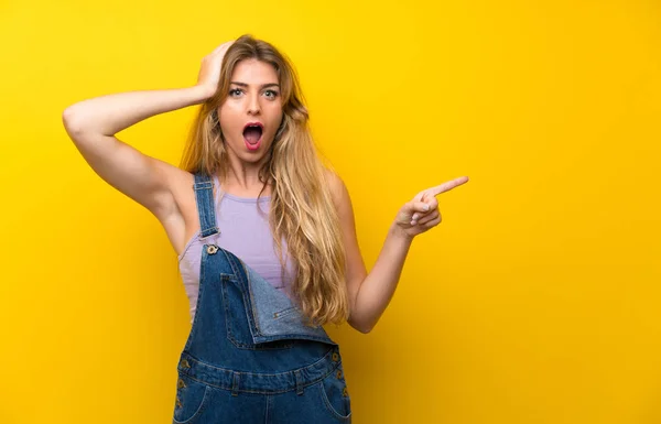 Young Blonde Woman Overalls Isolated Yellow Background Surprised Pointing Finger — Stock Photo, Image