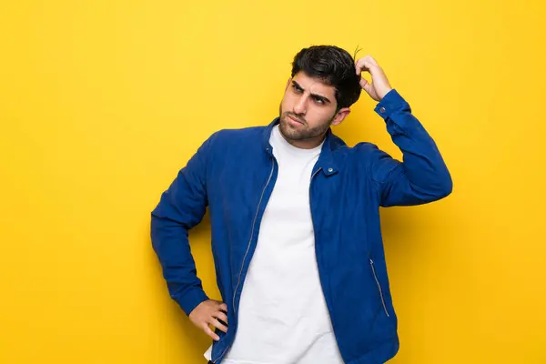 Man with blue jacket over yellow wall having doubts while scratching head