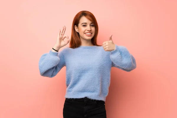 Young redhead woman over pink background showing ok sign with and giving a thumb up gesture