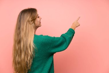 Pembe duvarın üstünde yeşil kazaklı genç sarışın kadın işaret parmağıyla işaret ediyor.