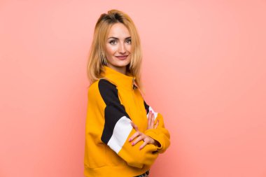 Sarışın, soyutlanmış, pembe arka planı olan, kollarını kavuşturup ileriye bakan genç bir kadın.