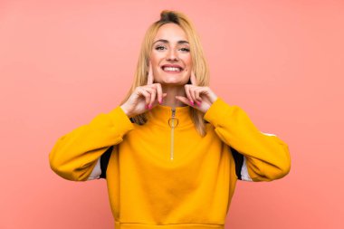 Mutlu ve hoş bir ifadeyle gülümseyen, soyutlanmış pembe arka planı olan genç sarışın kadın.