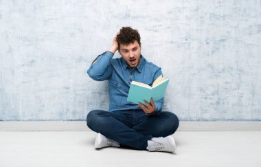 Genç adam yerde oturmuş bir kitap tutuyor ve okuyor.