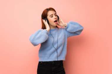 Pembe arka planda genç kızıl kadın kulaklıkla müzik dinliyor.