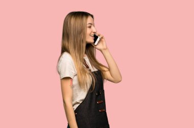 Young woman with apron keeping a conversation with the mobile phone on isolated pink background