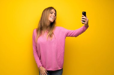 Sarı duvarın üstünde pembe kazaklı bir kadın selfie çekiyor.