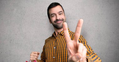 Alışveriş poşetleri ile Adam gülümseyen ve dokulu duvar üzerinde zafer işareti gösteren