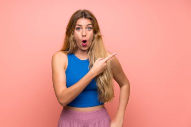 Soyutlanmış pembe arka planı olan genç sarışın kadın şaşırmış ve işaret ediyor.