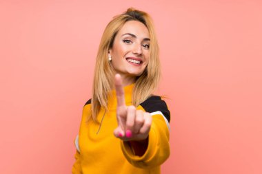 Soyutlanmış pembe arka planı olan genç sarışın kadın parmağını kaldırıyor ve gösteriyor.