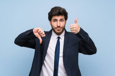 İzole edilmiş mavi duvarın üstündeki işadamı iyi-kötü işareti yapıyor. Evet ya da değil arasında kararsız