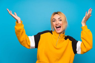 Soyutlanmış mavi arka planda, yüzünde sürpriz ifade olan genç sarışın kadın.