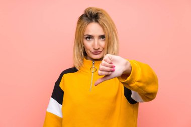 Soyutlanmış pembe arka planda genç sarışın kadın olumsuz bir ifadeyle baş parmağını aşağı gösteriyor.