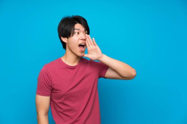 Izole mavi duvar üzerinde kırmızı gömlek li Asyalı adam ağzı geniş açık bağırarak