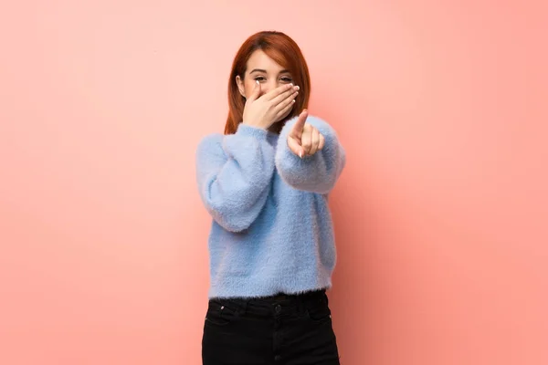 Young redhead woman over pink background pointing with finger at someone and laughing
