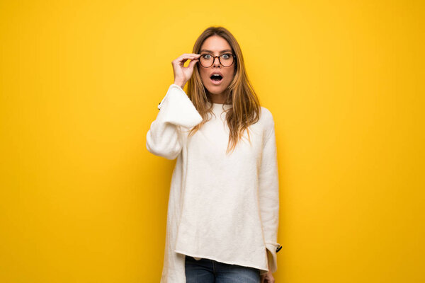 Blonde woman over yellow wall with glasses and surprised