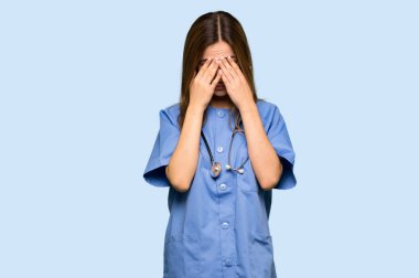 İzole edilmiş mavi arka planda yorgun ve hasta suratlı genç hemşire.