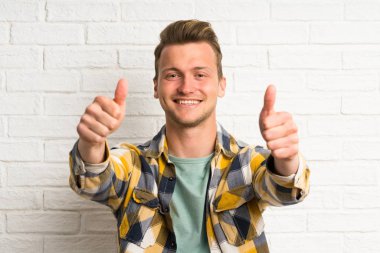 Beyaz tuğla duvar üzerinde Sarışın yakışıklı adam jest kadar bir başparmak veren