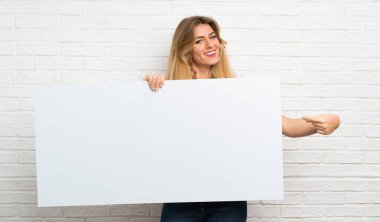Beyaz tuğlalı duvarın üzerinden geçen genç sarışın kadın bir konsept eklemek için boş beyaz bir levha tutuyor.