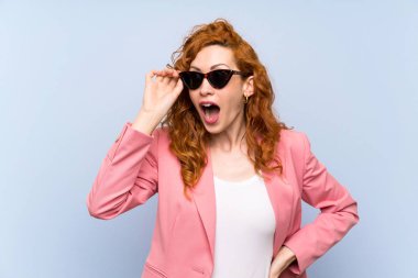Gözlüklü izole mavi duvar üzerinde takım elbiseli Redhead kadın ve şaşırttı