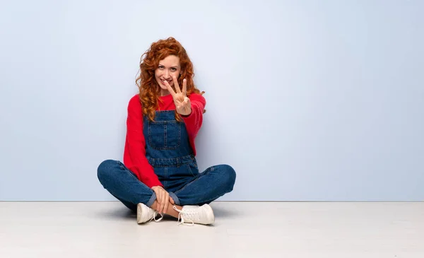 Redhead woman with overalls sitting on the floor happy and counting three with fingers