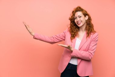 İzole pembe duvar üzerinde takım elbiseli Redhead kadın gelmeye davet için yan ellerini uzatıyor