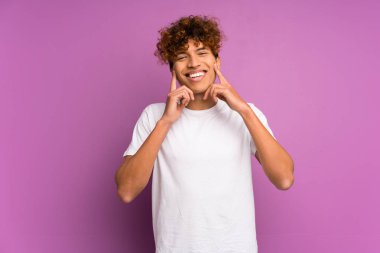 İzole edilmiş mor duvarın ardında mutlu ve hoş bir ifadeyle gülümseyen genç bir Afrikalı Amerikalı.