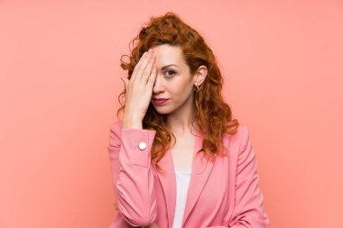 Elle bir göz kapsayan izole pembe duvar üzerinde takım elbiseli Redhead kadın