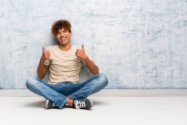 Genç Afro-Amerikan bir adam yerde oturuyor. Başparmakları havada ve gülümsüyor.