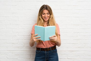 Beyaz tuğlalı duvarın üzerinde elinde kitap tutan ve okuyan genç sarışın kadın.