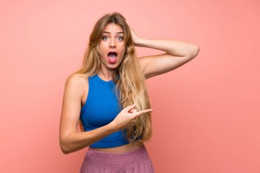 Soyutlanmış pembe arka planı olan genç sarışın kadın şaşırdı ve işaret etti.