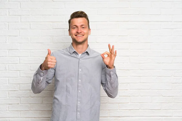 Blondynka Człowiek Przez Biały Cegła Ściana Pokazując Znak Kciuk Górę — Zdjęcie stockowe