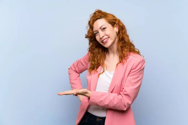 Redhead woman in suit over isolated blue wall presenting an idea while looking smiling towards