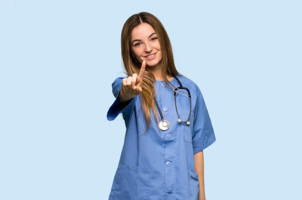 Ung Sjuksköterska Visar Och Lyfter Ett Finger Isolerad Blå Bakgrund — Stockfoto