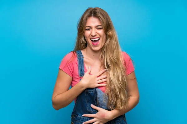 Young blonde woman with overalls over isolated blue wall smiling a lot