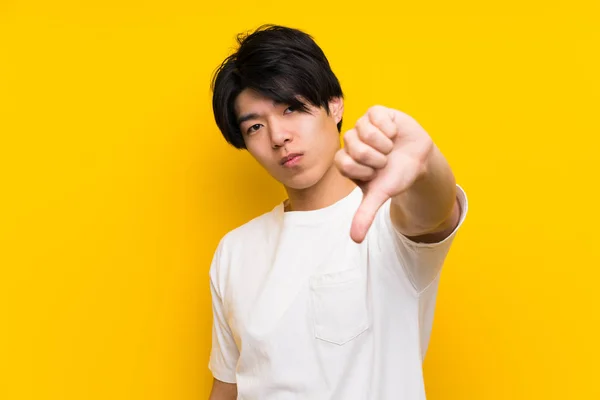 孤立した黄色い壁の上のアジア人男性は 否定的な表情で親指を下に示す — ストック写真