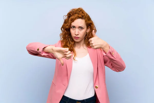 Redhead woman in suit over isolated blue wall making good-bad sign. Undecided between yes or not