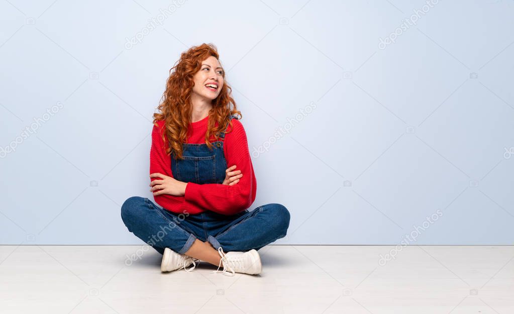 Redhead woman with overalls sitting on the floor with arms crossed and happy