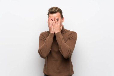 Elleriyle gözleri kaplayan izole beyaz duvar üzerinde Blonde adam ve parmaklardan bakarak