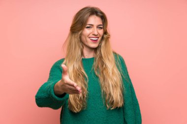 Pembe duvarın üstünde yeşil kazaklı genç sarışın kadın el sıkışıyor.