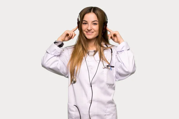 Young doctor woman listening to music with headphones on isolated grey background