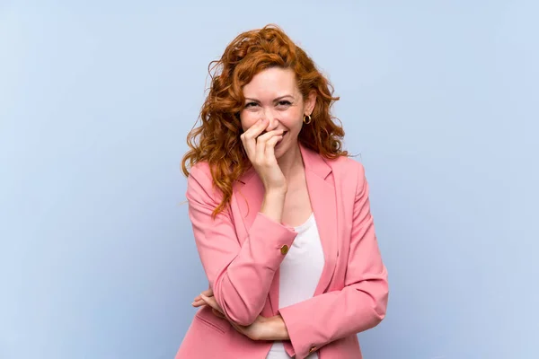 Redhead woman in suit over isolated blue wall smiling a lot