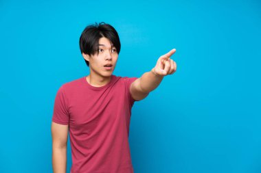 Izole mavi duvar üzerinde kırmızı gömlek ile Asyalı adam uzağa işaret