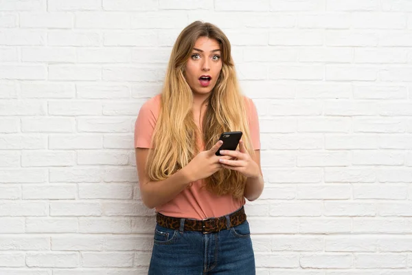 年轻的金发女子用手机翻过白砖墙 — 图库照片