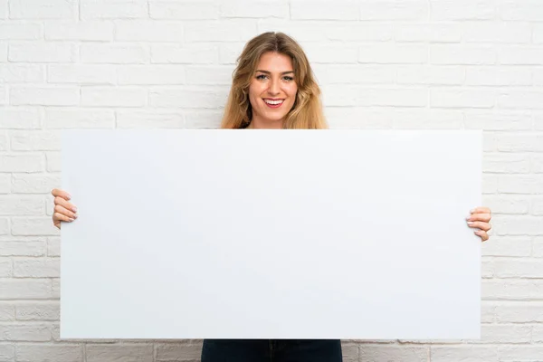 白砖墙上的金发年轻女子拿着一个空的白色标语牌插入一个概念 — 图库照片